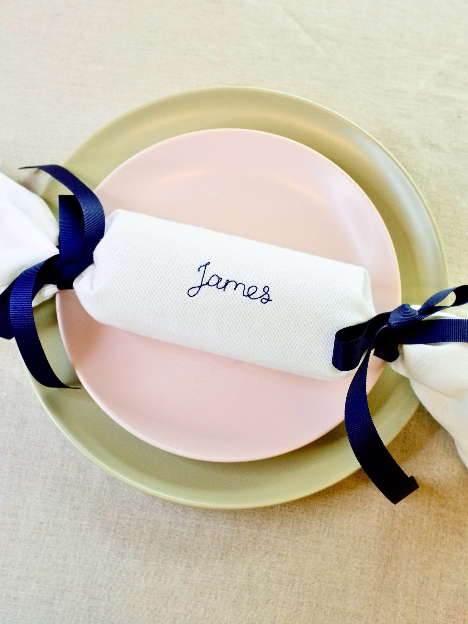 Single personalised reusable cracker napkin embroidered with the name James in navy thread, tied with navy ribbons. Fabric Christmas cracker alternative for sustainable holiday dining.