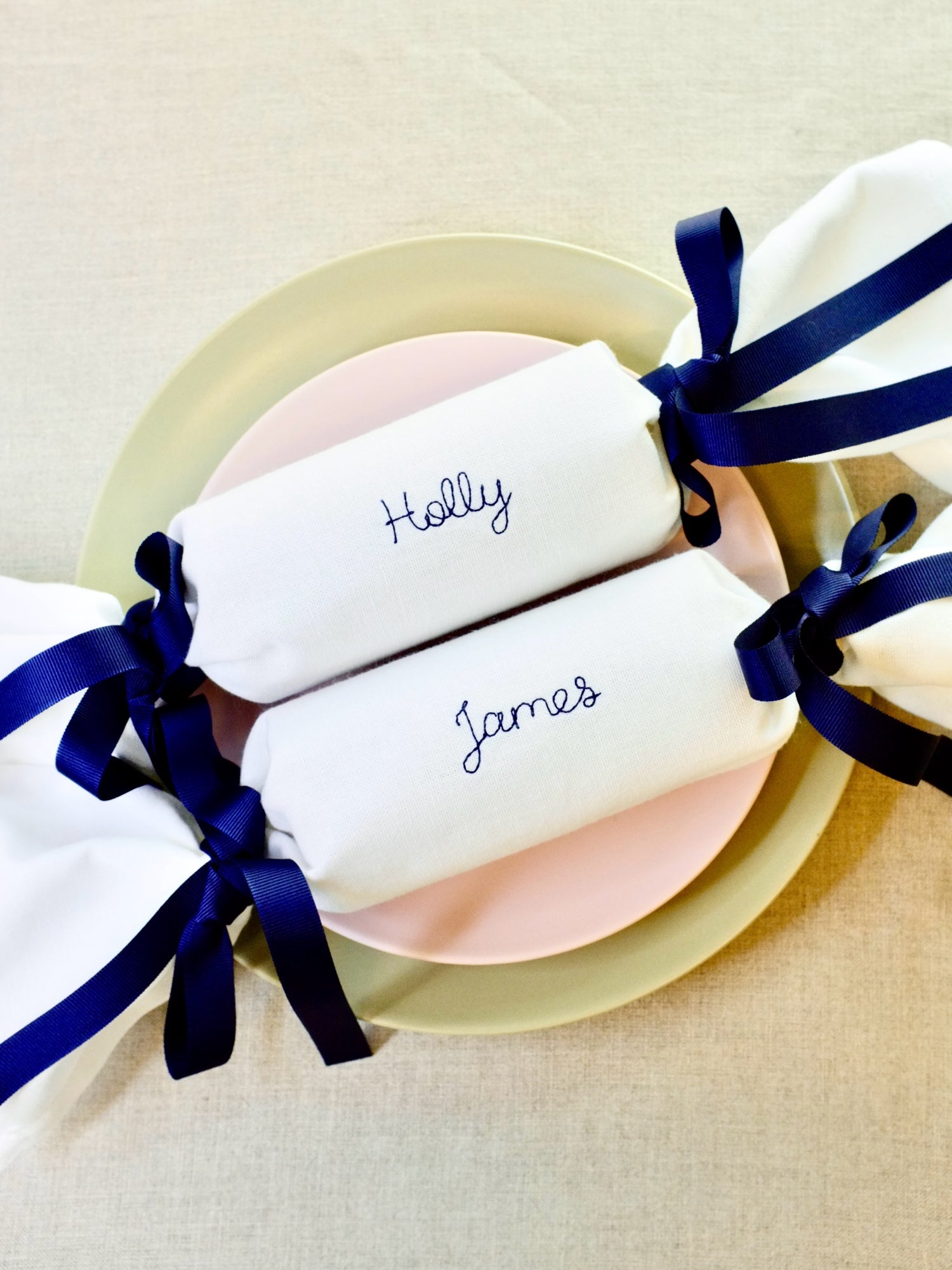 Reusable embroidered fabric Christmas crackers with names Holly and James stitched in navy thread, tied with matching navy ribbons. Personalised napkin crackers for eco-friendly festive tables.