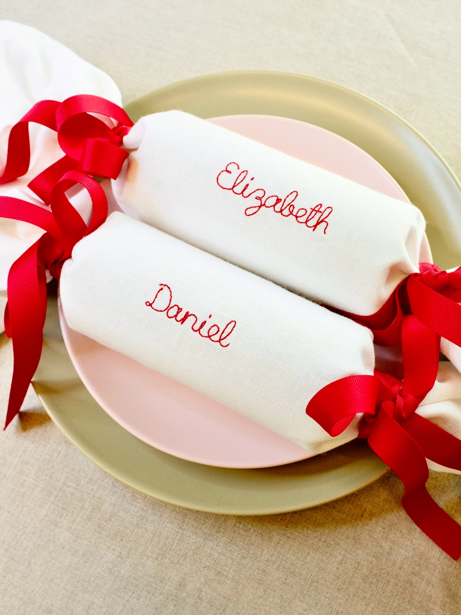 Set of personalised embroidered Christmas cracker napkins with names Elizabeth and Daniel in red thread, finished with red ribbon, displayed on layered plates. Sustainable, reusable fabric crackers.