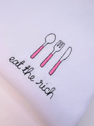 White tshirt with embroidered pink cutlery and embroidery eat the rich  font