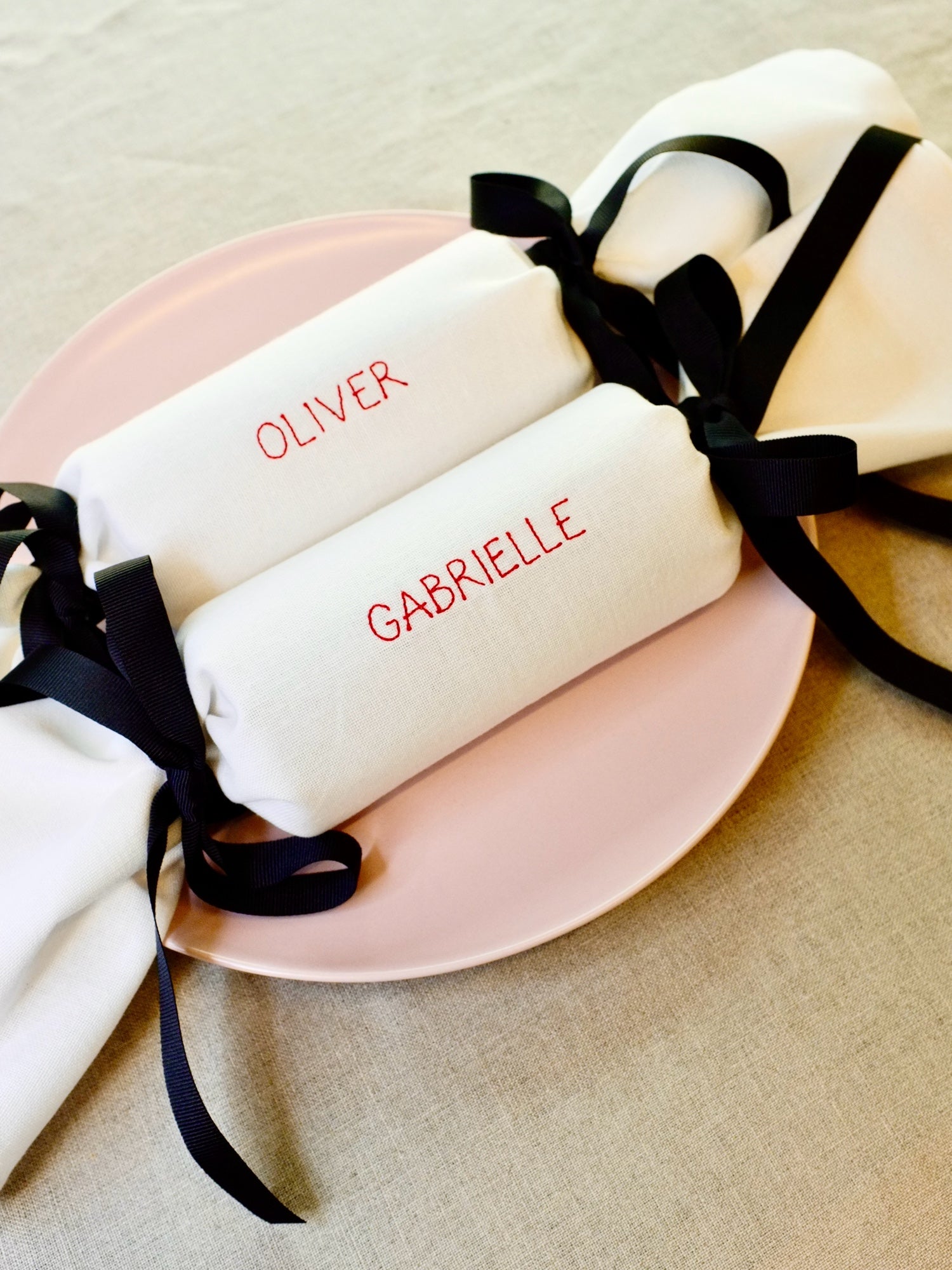 Personalised reusable fabric Christmas crackers embroidered with names Oliver and Gabrielle in red thread, tied with black ribbon, styled on a pink plate. Perfect eco-friendly alternative to disposable crackers.