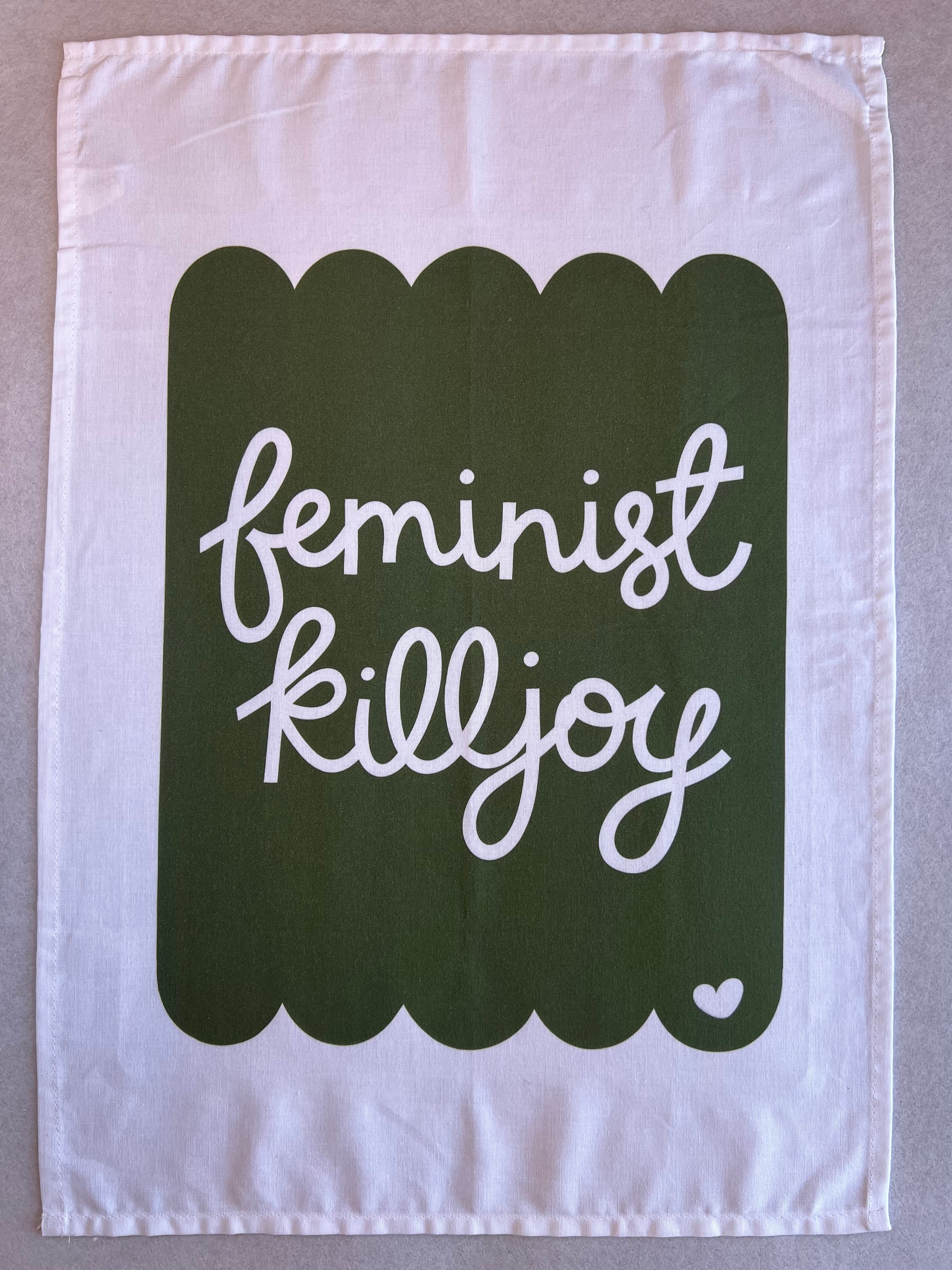 Cotton tea towel with the phrase ‘feminist killjoy’ in bold white script on a soft pink background with a scalloped border. The towel features a small heart detail in the corner