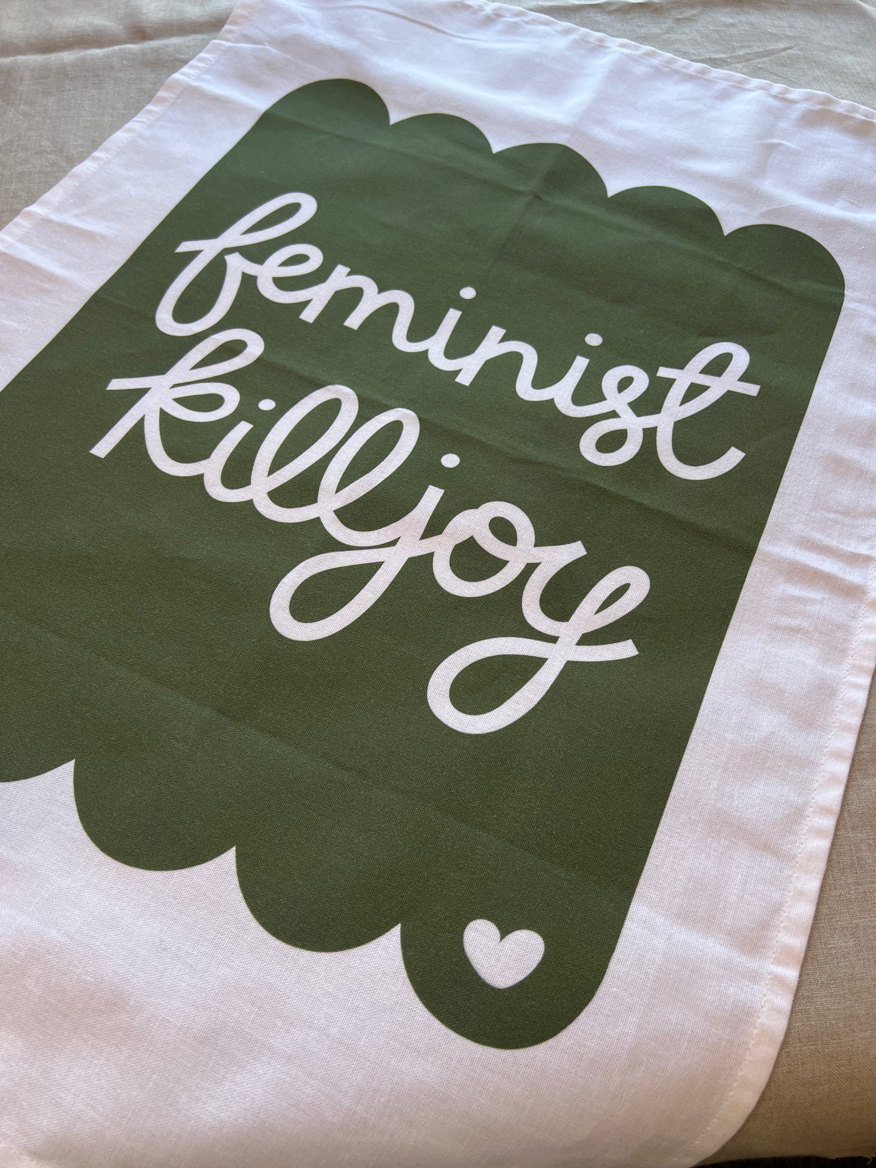 Cotton tea towel with the phrase ‘feminist killjoy’ in bold white script on a soft pink background with a scalloped border. The towel features a small heart detail in the corner
