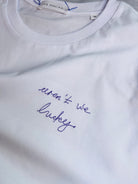 White tee with the phrase ‘aren’t we lucky’ embroidered handwriting in purple thread 