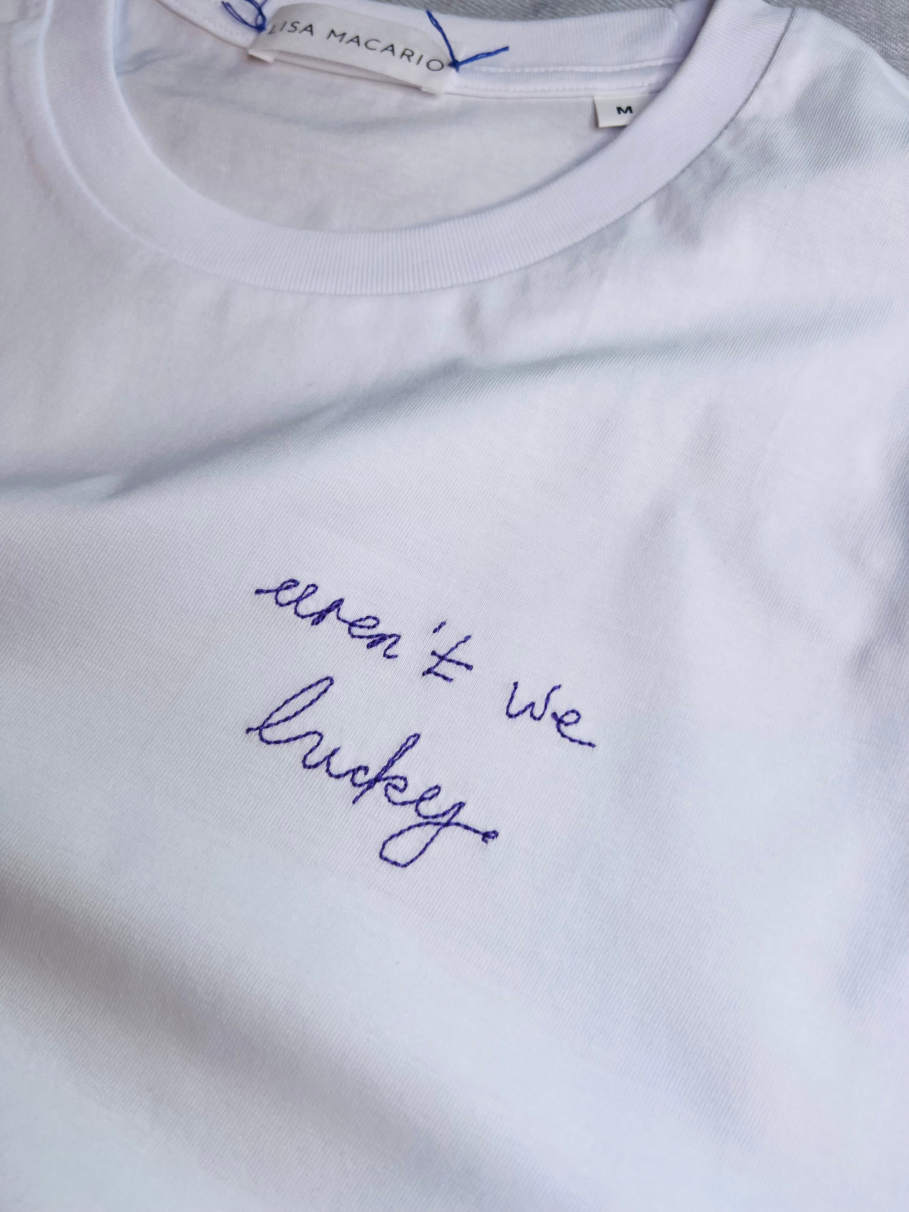 White tee with the phrase ‘aren’t we lucky’ embroidered handwriting in purple thread 