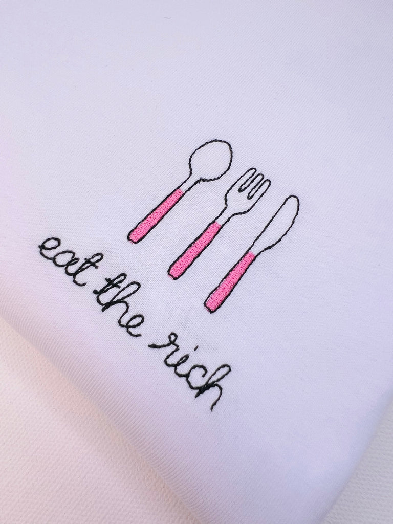 White tshirt with embroidered pink cutlery and embroidery eat the rich  font 